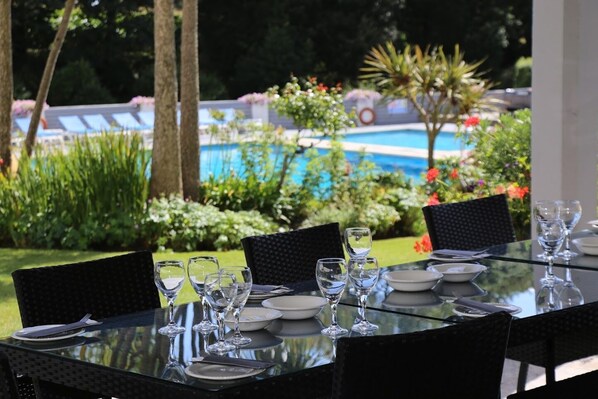 Una mesa de comedor al aire libre con sillas, preparada para una comida, con vista a una piscina y un jardín exuberante.