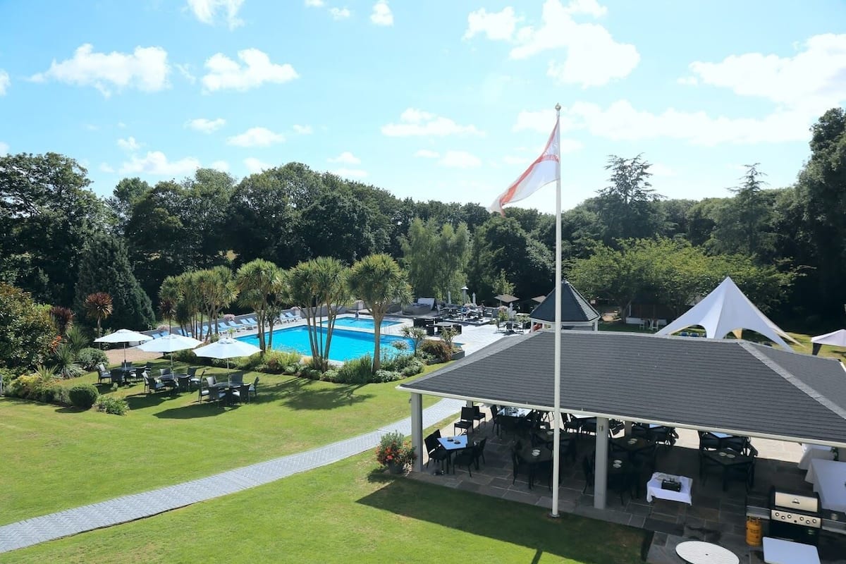 A pool area with lounge chairs and umbrellas, surrounded by trees and a flagpole.