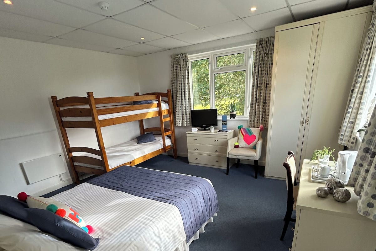 A bedroom with a bunk bed, a desk with a computer, a chair, a wardrobe, and a window with curtains.
