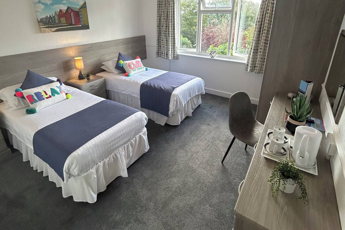 A hotel room with two beds, a desk, a chair, a window with curtains, a painting on the wall, and a small plant on the desk.