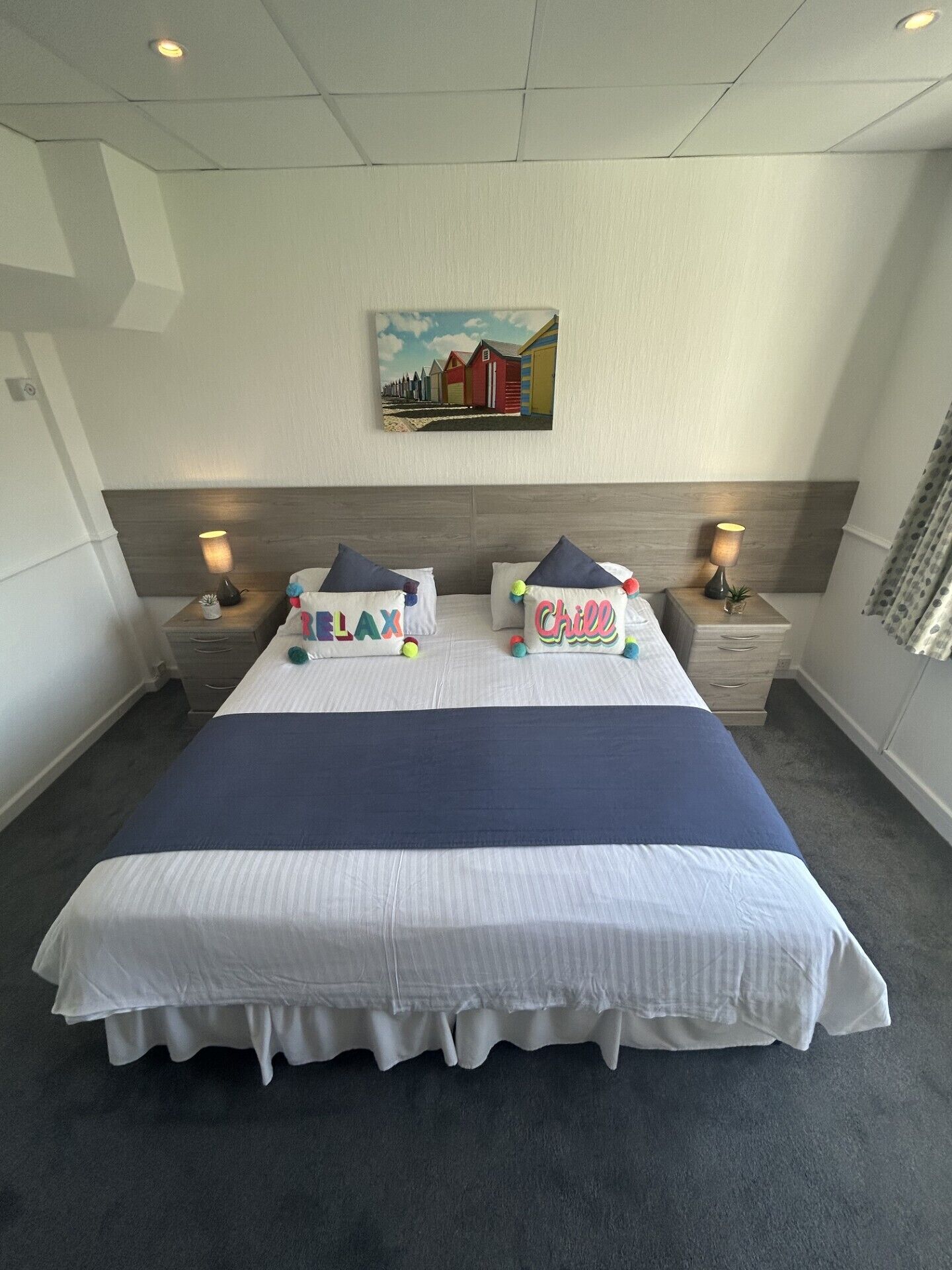 A neatly made bed with a blue bedspread and white bedding, two bedside tables with lamps, a painting on the wall, and a grey carpet.