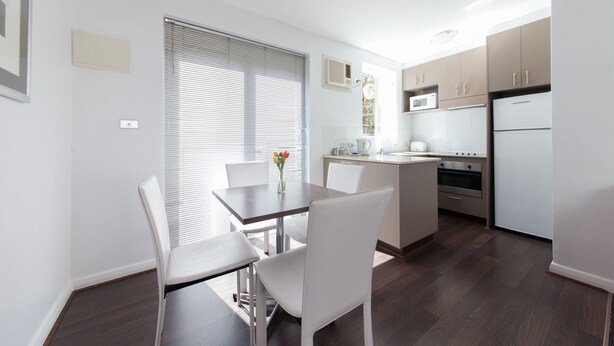 A modern kitchen with white cabinets, a dining table with chairs, and a window with blinds. - Easystay Apartments (St. Kilda East)