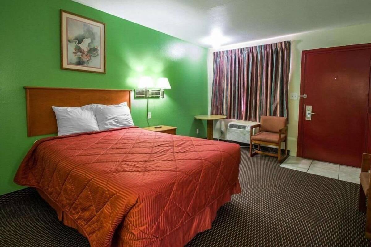 A hotel room with a bed, a chair, a table, a lamp, and a door.