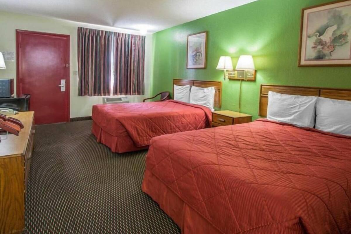 A hotel room with two beds, a desk, a chair, a door, a window with curtains, and wall-mounted lamps.