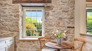 A stone wall interior with a window, a round wooden table, two wicker chairs, and a vase with flowers.
