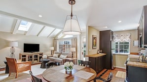 A spacious living area with a large round table, comfortable seating, a flat-screen TV, and a kitchen area with a window.