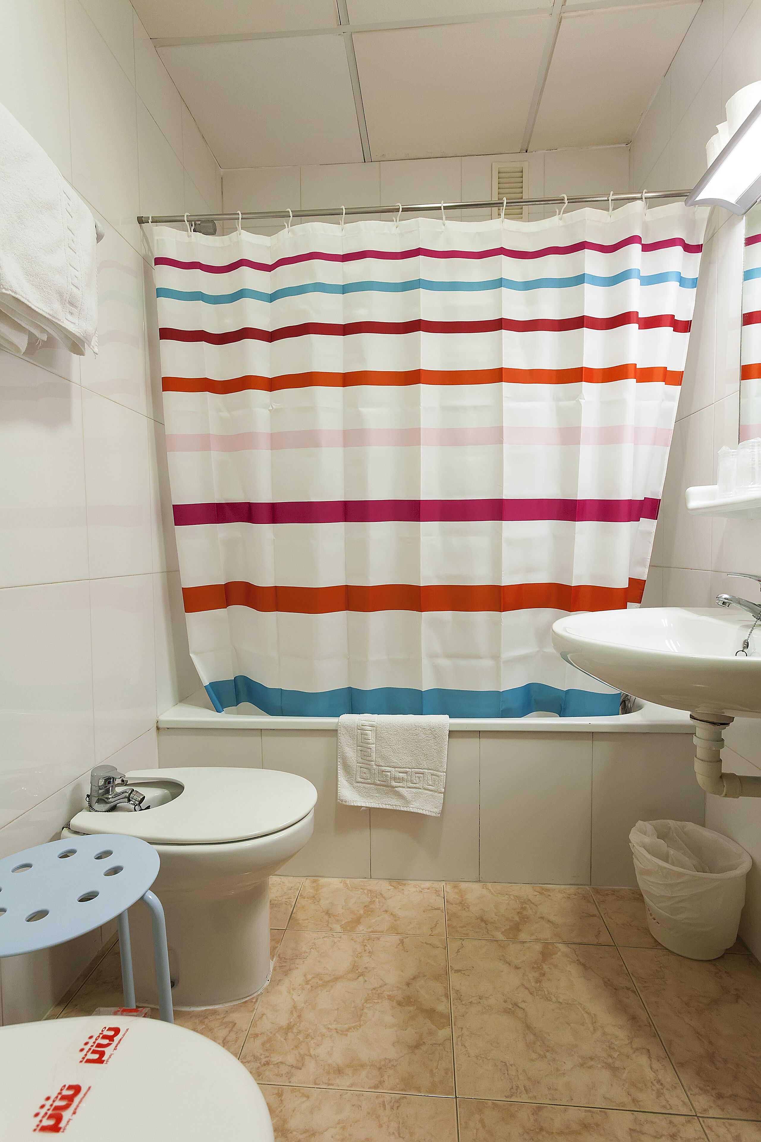 Un baño con cortina de ducha a rayas, un inodoro, un lavabo y un taburete pequeño.