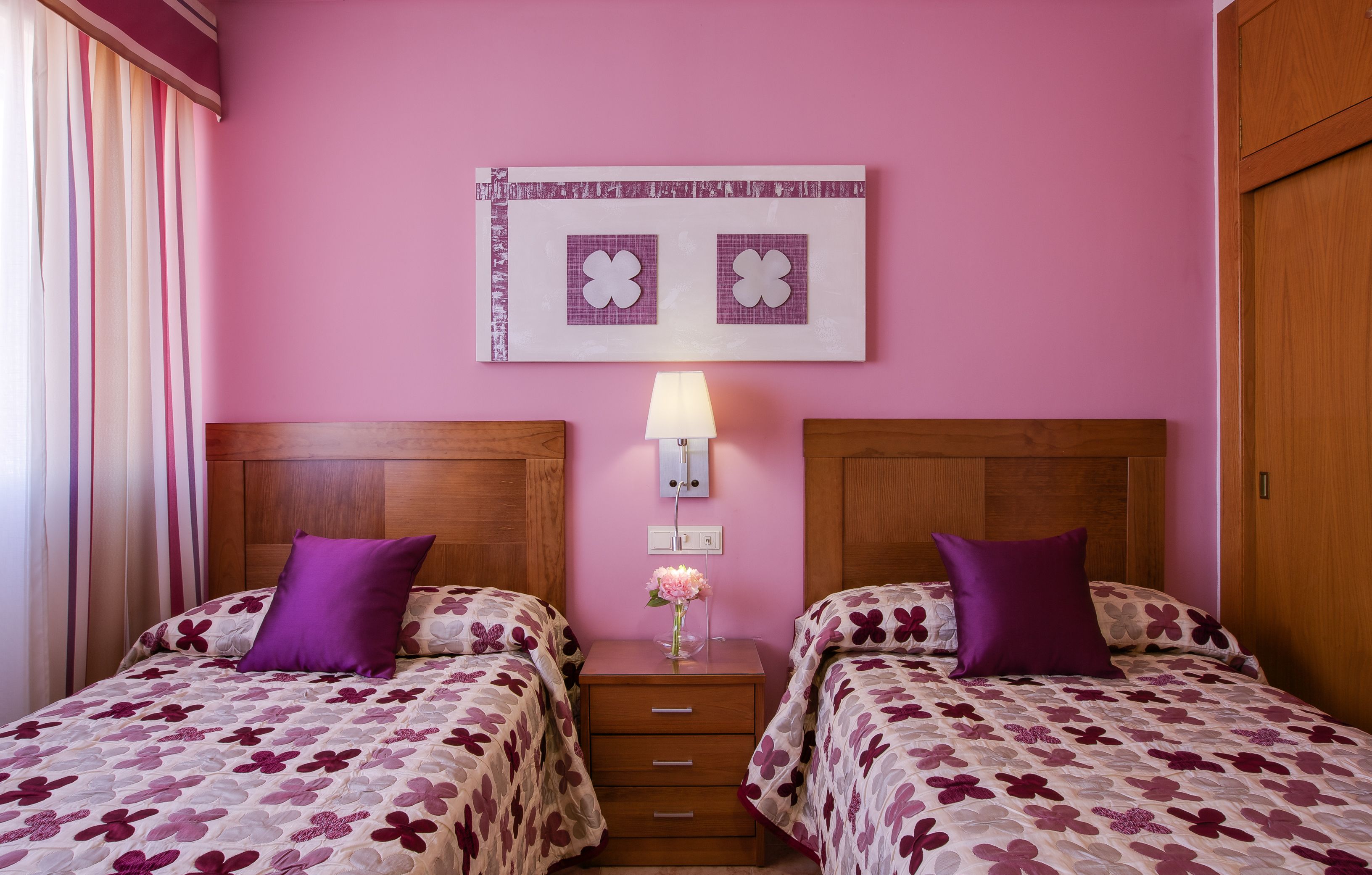 Une chambre d’hôtel avec deux lits, chacun recouvert d’une couvre-lit à motifs floraux violets et agrémenté d’oreillers assortis. Une table de chevet avec une lampe et un vase de fleurs se trouve entre les deux lits. Le mur est rose, et une œuvre d’art encadrée est accrochée au-dessus de la table de chevet.