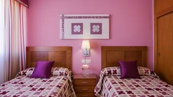 Une chambre d’hôtel avec deux lits, chacun recouvert d’une couvre-lit à motifs floraux violets et agrémenté d’oreillers assortis. Une table de chevet avec une lampe et un vase de fleurs se trouve entre les deux lits. Le mur est rose, et une œuvre d’art encadrée est accrochée au-dessus de la table de chevet.