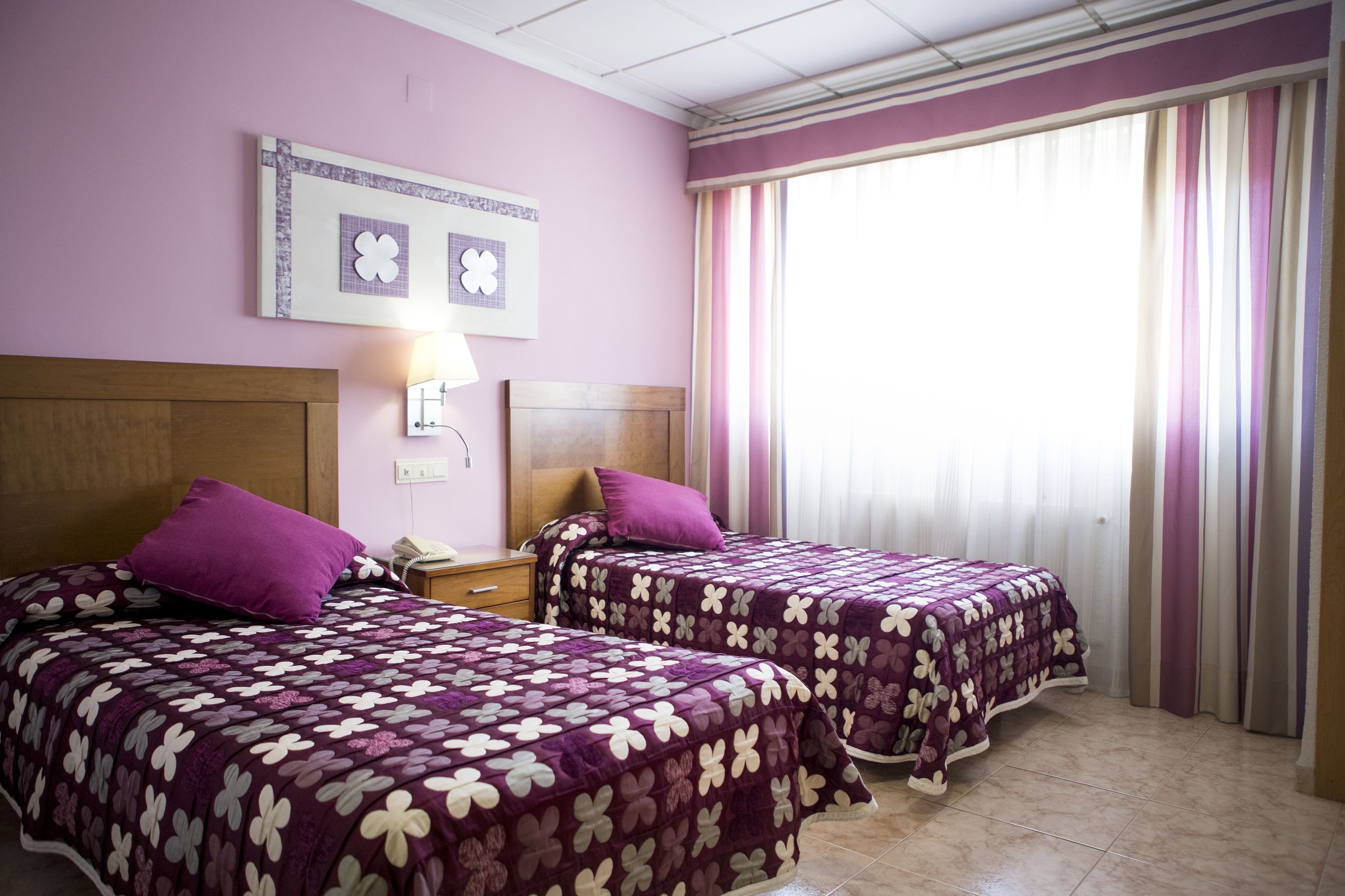 A hotel room with two beds, each with a purple floral bedspread, a wooden headboard, a bedside table with a lamp, and a window with striped curtains.