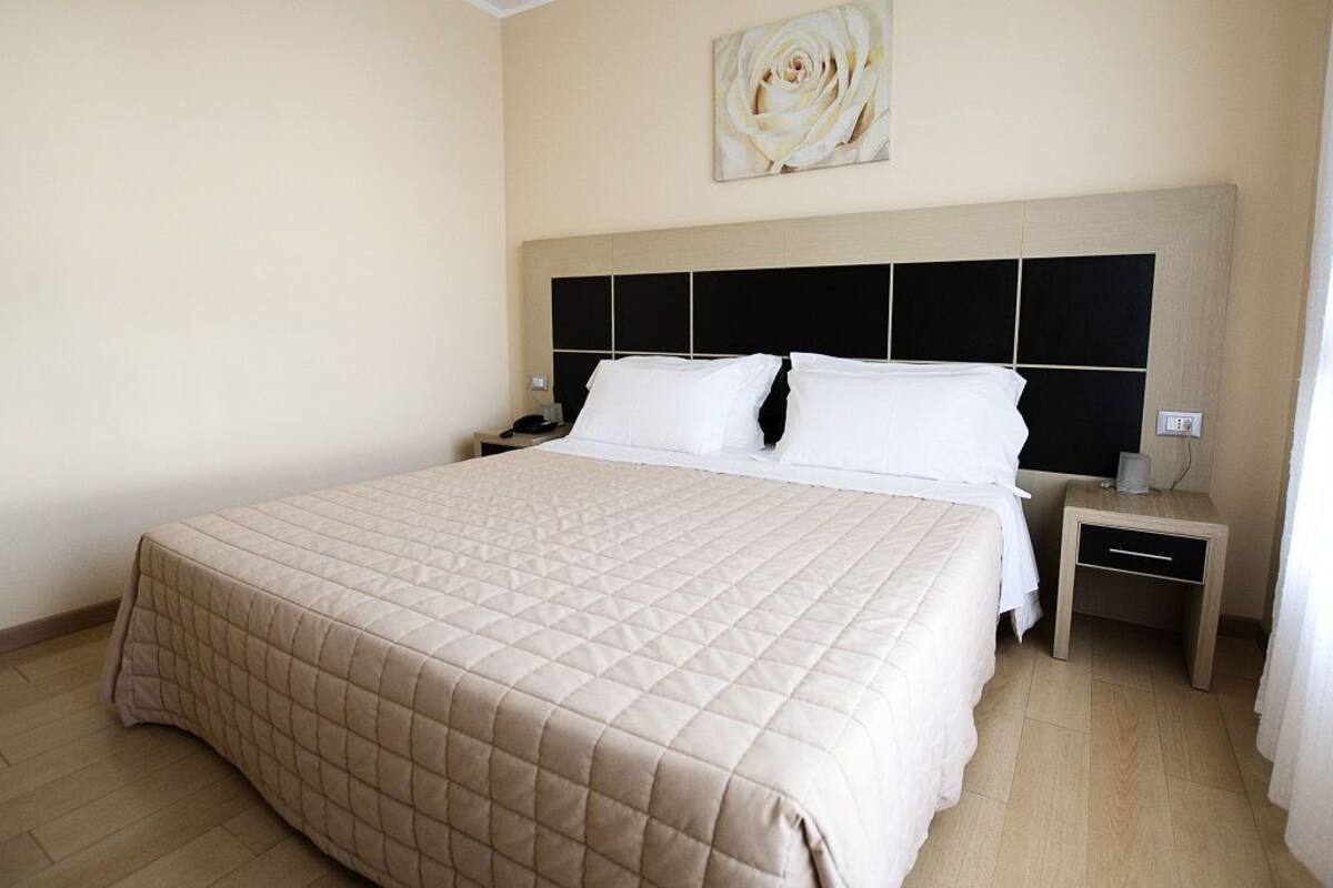 A hotel room with a large bed, a headboard with a black panel design, a bedside table with a lamp, and a wall-mounted artwork.