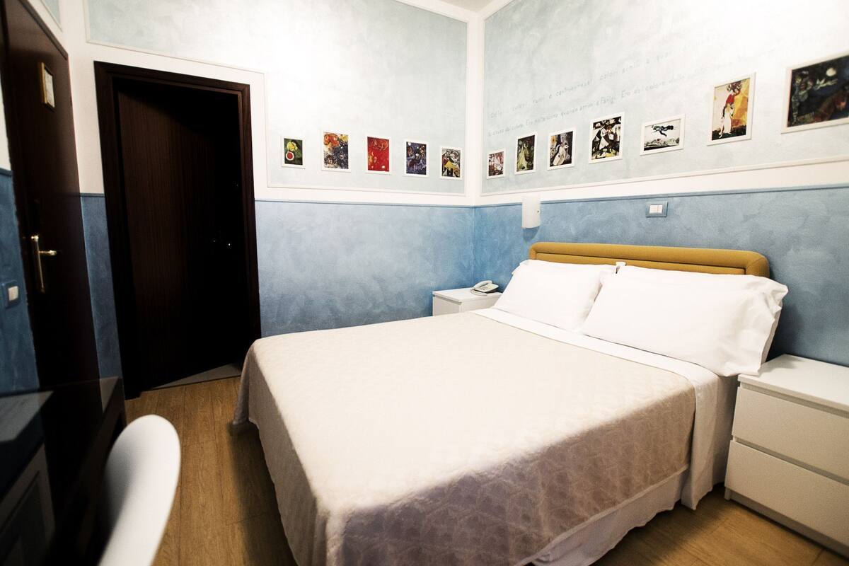 A bedroom with a bed, bedside tables, a desk, and a chair. There are wall-mounted pictures and a door.