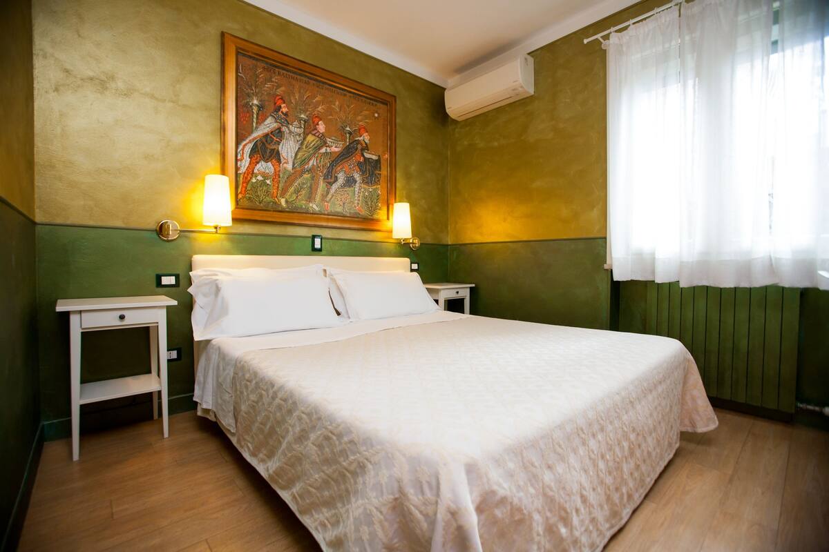 A hotel room with a large bed, two bedside tables, a wall-mounted painting, and a window with sheer curtains.