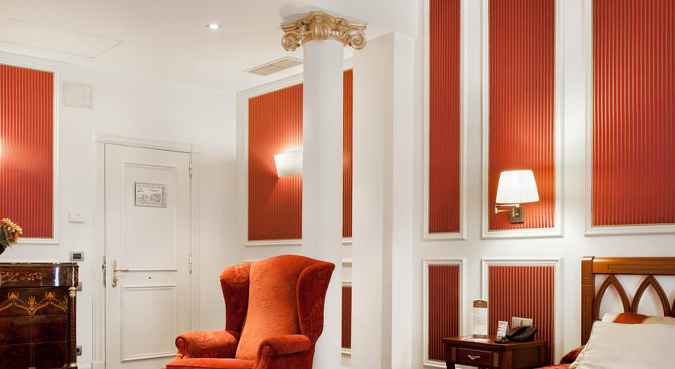 Habitación de hotel con una amplia butaca tapizada de rojo, una mesita de noche de madera con lámpara y una cama con cabecero de madera.