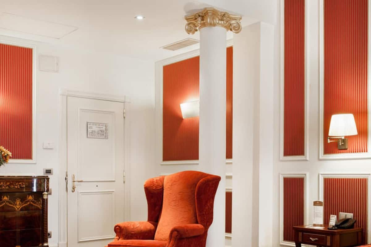 Habitación de hotel con una amplia butaca tapizada de rojo, una mesita de noche de madera con lámpara y una cama con cabecero de madera.