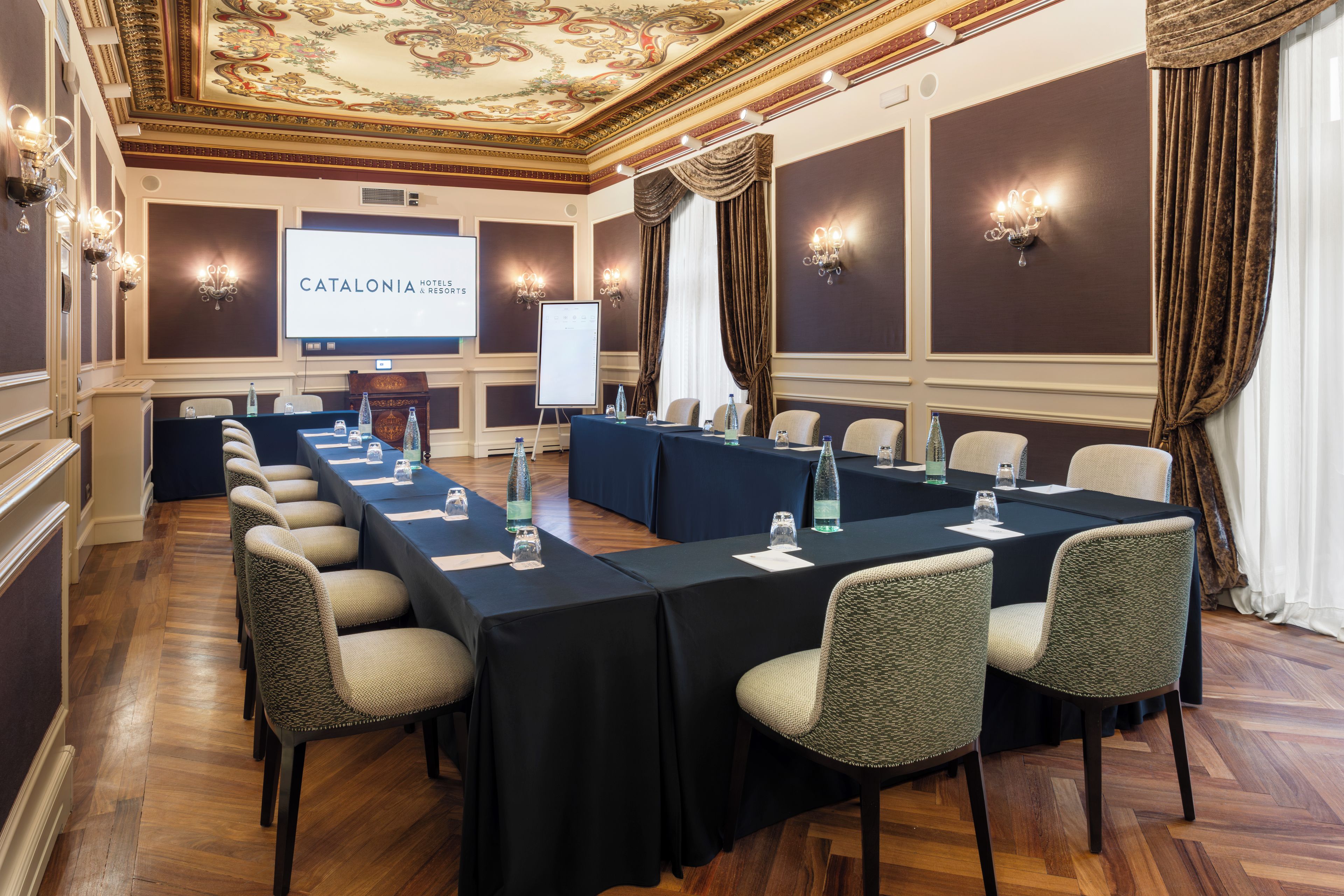 A conference room with a long table, chairs, a projector screen, and a presentation about Catalonia Hotels & Resorts.