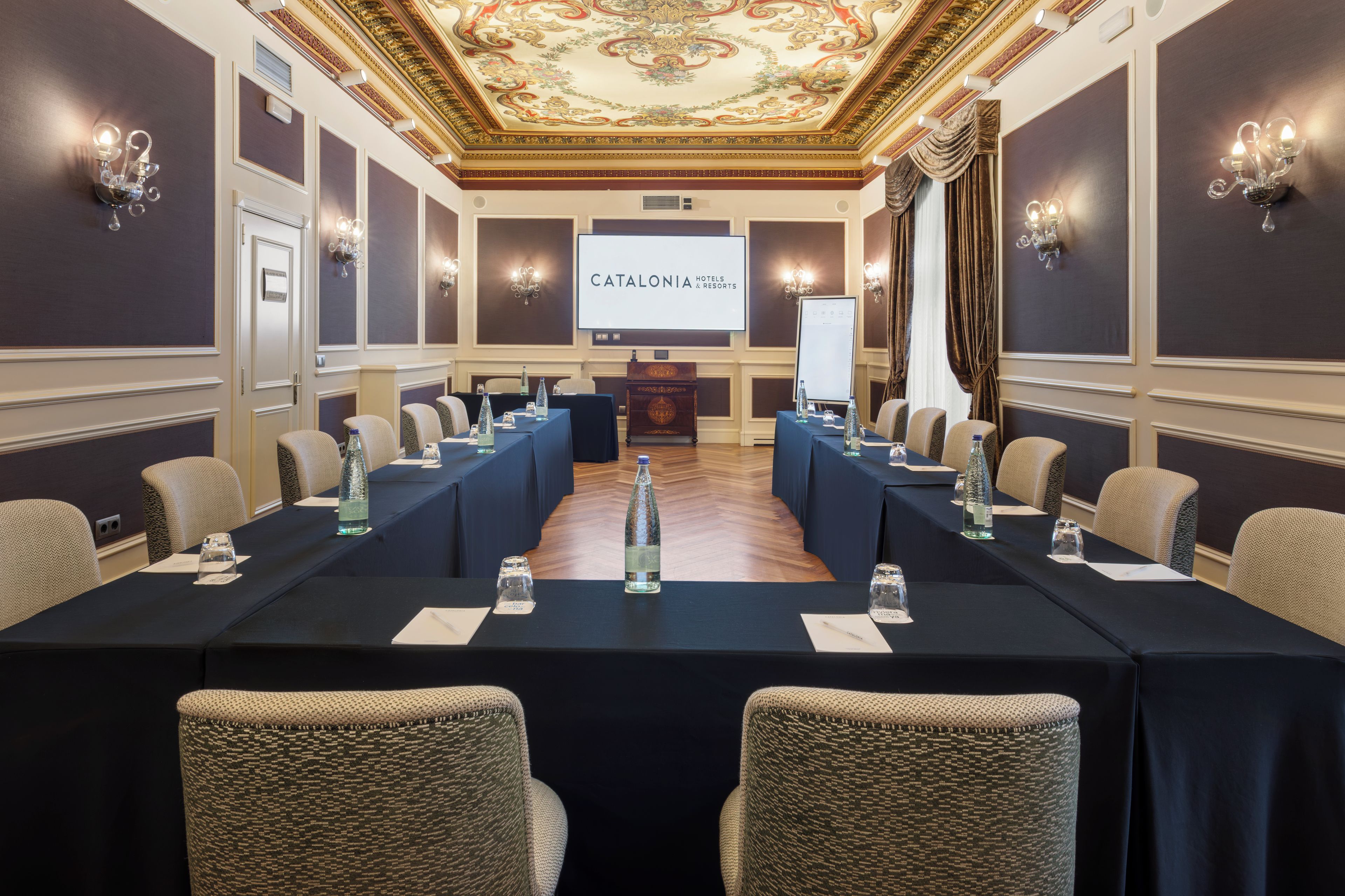 Un salón de conferencias con una mesa larga, sillas y una pantalla mostrando 'CATALONIA HOTELS & RESORTS'.