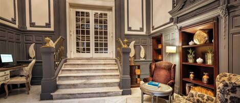 A grand staircase leading to a door, a plush armchair, a small table with books, and a bookshelf with decorative items.