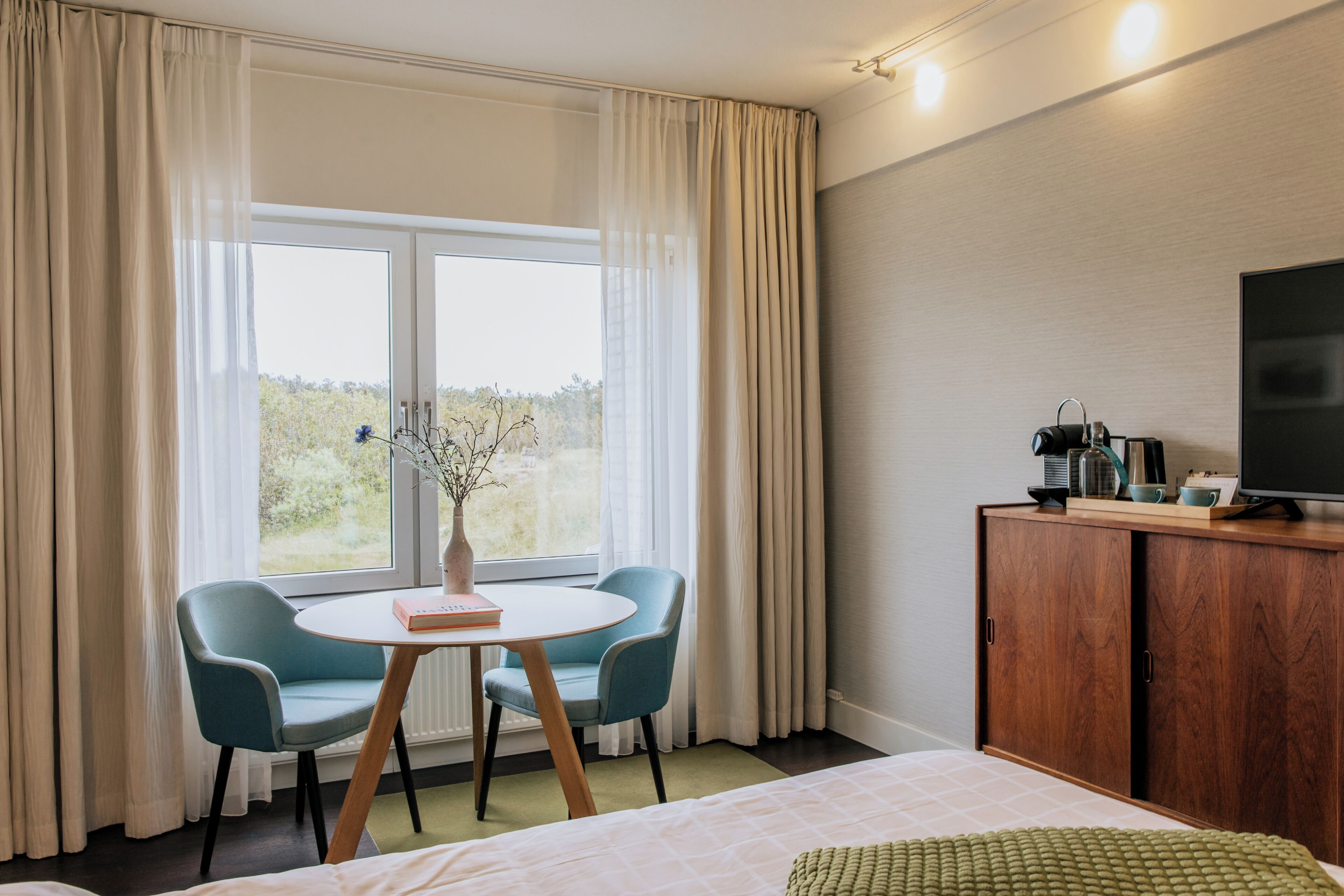 Une chambre d’hôtel comprenant un lit, une télévision, une armoire en bois, une table avec des chaises et une fenêtre avec des rideaux.