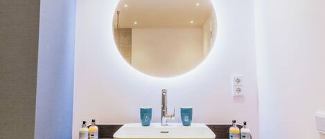 A bathroom with a rectangular sink, a round mirror, and two blue cups on the counter.