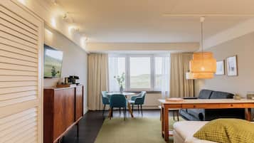 Quarto de hotel moderno com área de refeições, sofá e uma grande janela com cortinas.