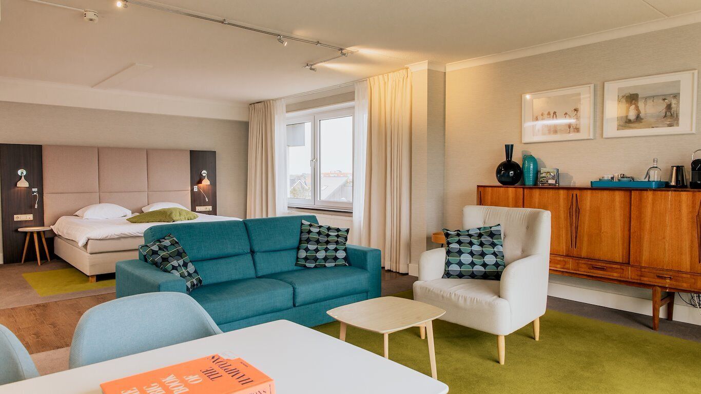 Ein modernes Hotelzimmer mit einem Bett, einem blauen Sofa, einem weißen Sessel, einem kleinen Tisch und einem Holzkabinett.