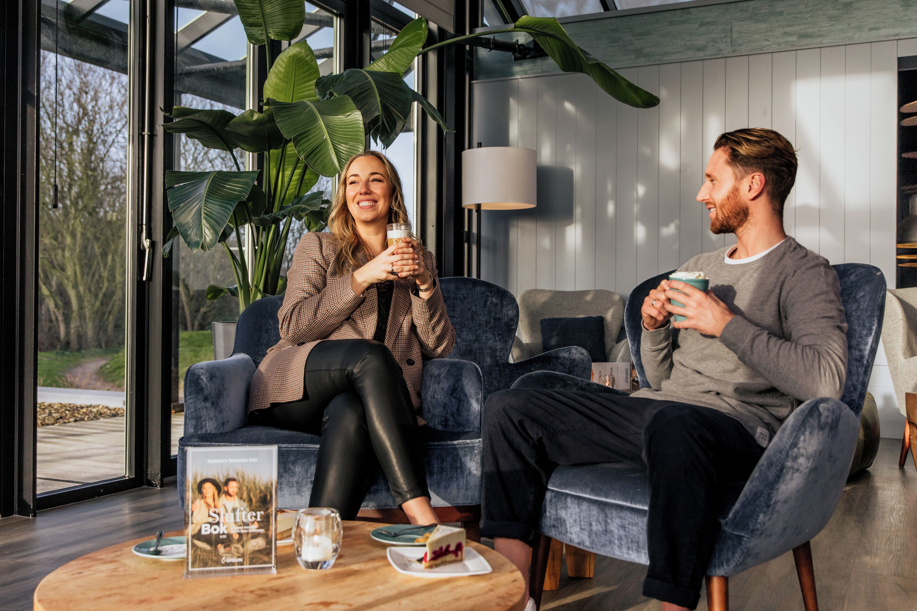 En moderne stue med to personer siddende på en blå sofa, et sofabord med kage og et indrammet foto, og en stor plante i baggrunden.