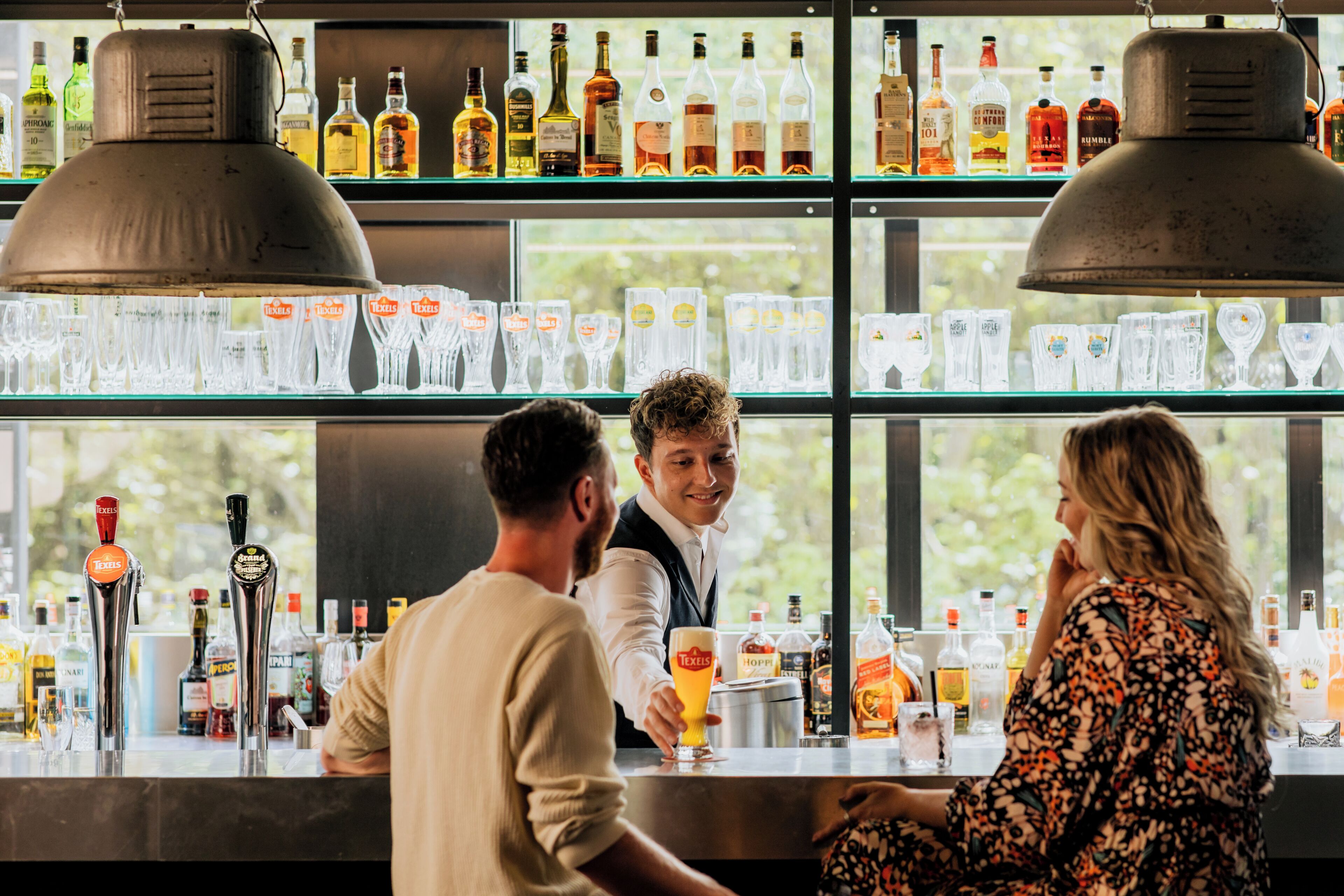 A bar with multiple liquor bottles and glasses on shelves, a bartender serving a customer, and a window view.