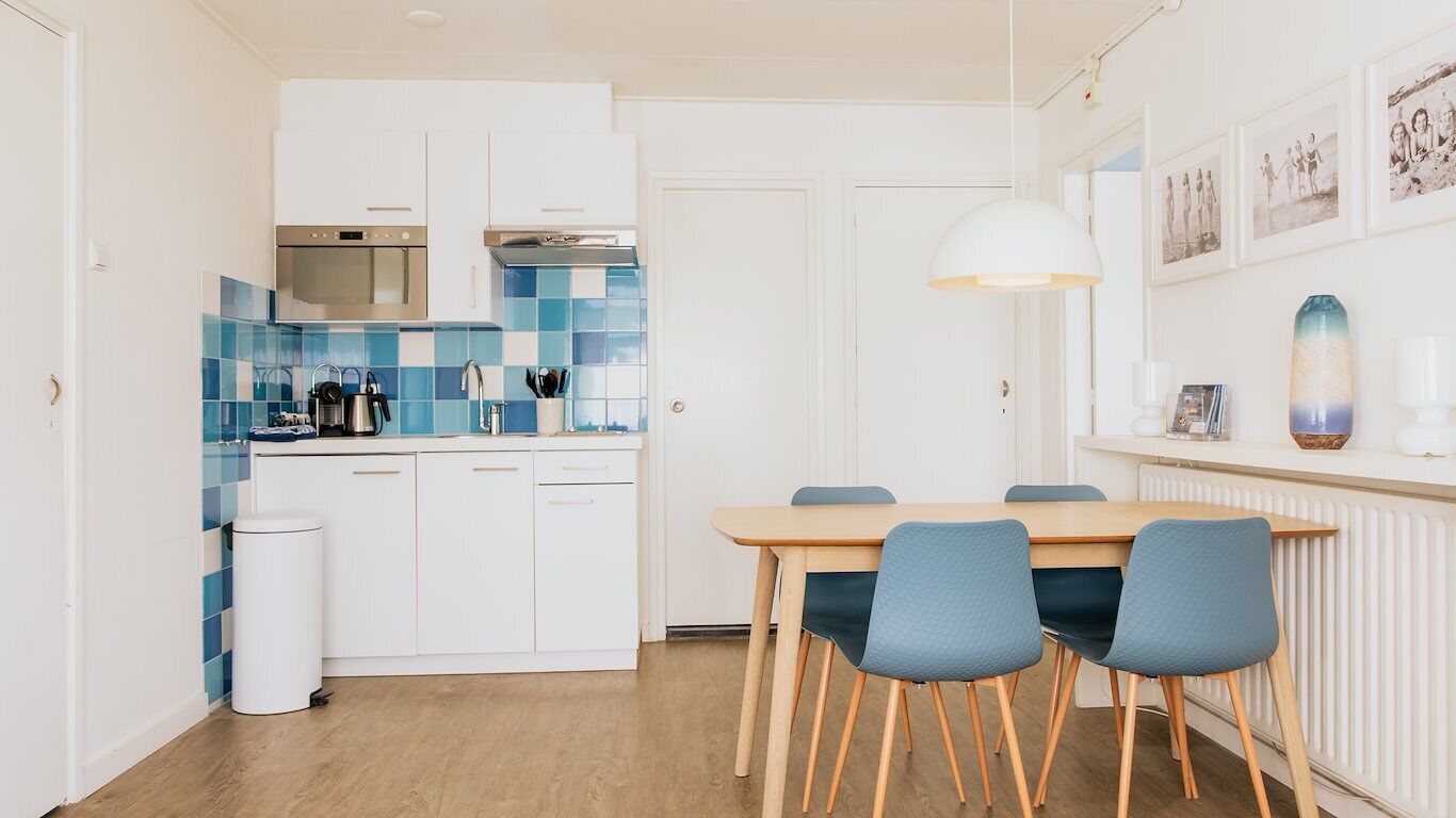 A modern kitchen with white cabinets, blue and white tiles, a wooden dining table with blue chairs, and a white pendant light.