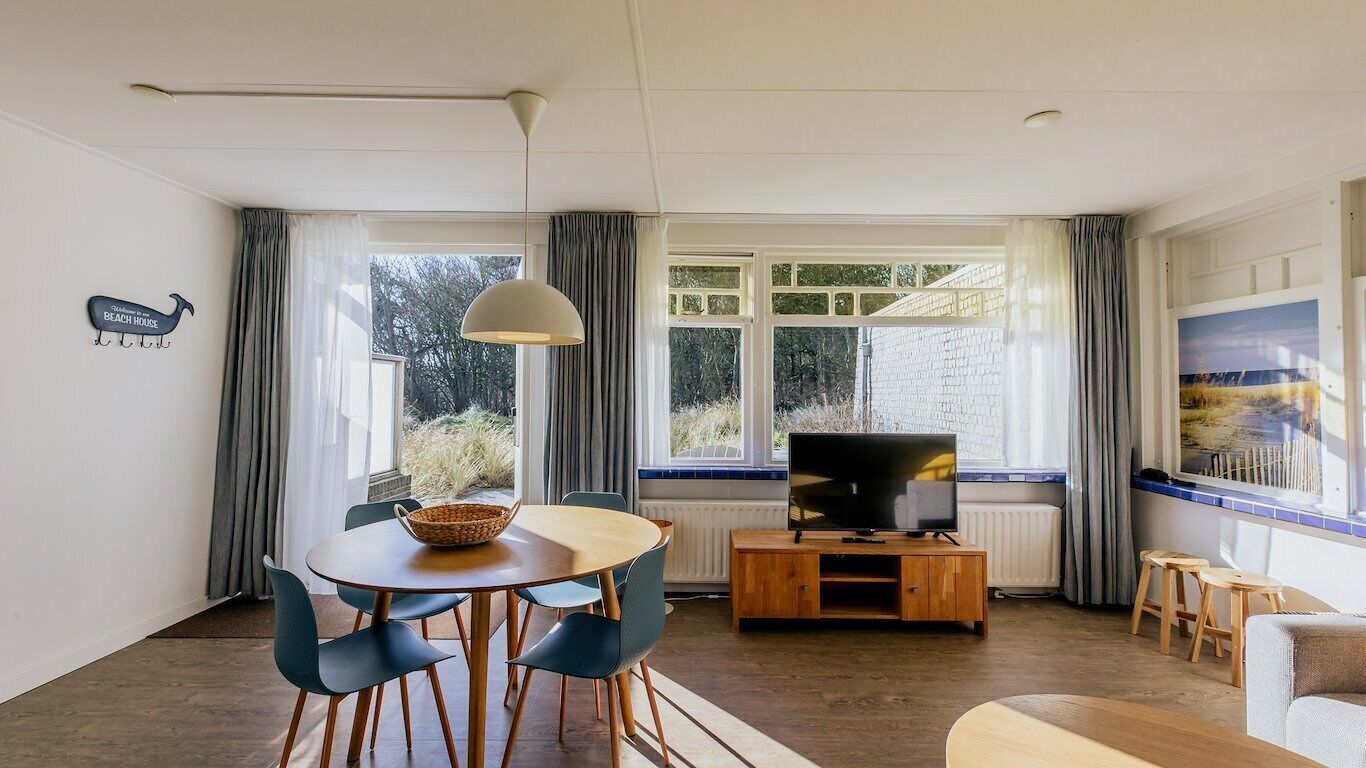 Una sala de estar con una mesa redonda de madera, sillas azules, un televisor sobre un mueble de madera y un amplio ventanal con cortinas.