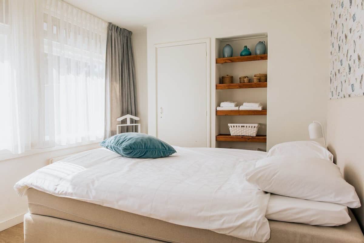 Een slaapkamer met een bed, een raam met dunne gordijnen, een inbouwkast met opbergmanden en een wandlamp.