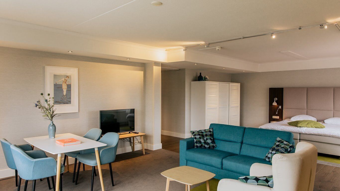 A modern living room with a teal sofa, a dining table with chairs, a TV, and a bed with a headboard.
