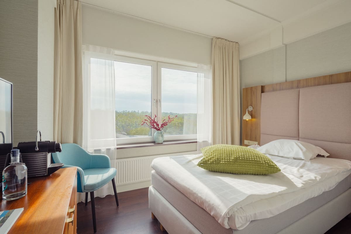 Een hotelkamer met een bed, een bureau met stoel, een raam met gordijnen en een vaas met bloemen op de vensterbank.