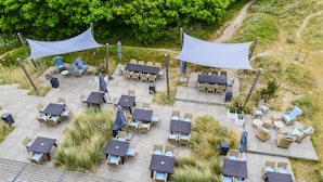 Une terrasse extérieure dotée de plusieurs tables et chaises, entourée de verdure et bordée d’un sentier pédestre.