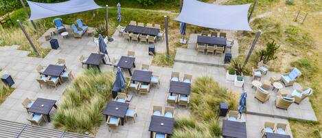 An outdoor patio with multiple tables and chairs, surrounded by greenery and a walking path.