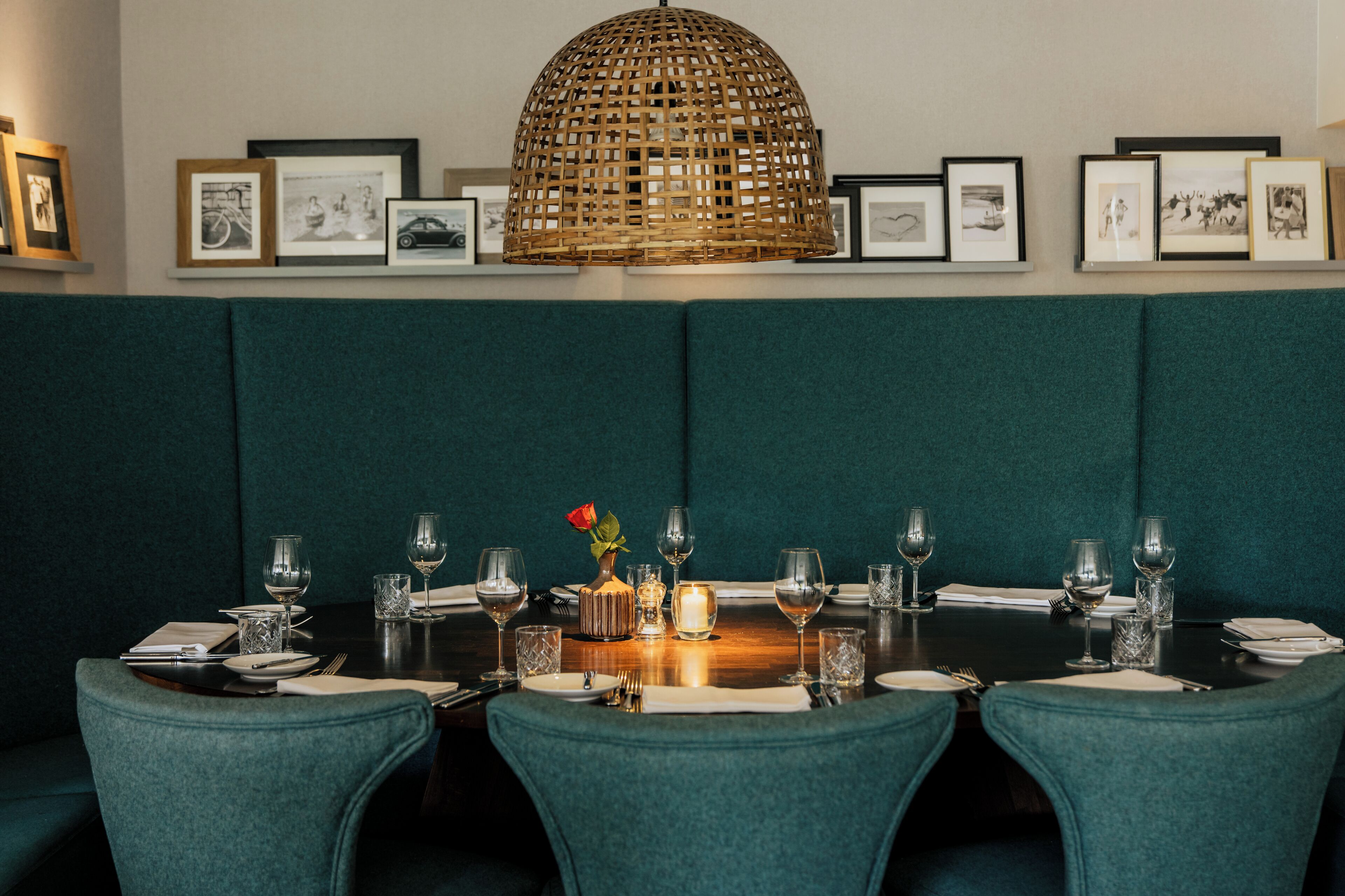 A dining table set for six with a candle centerpiece, framed pictures on the wall, and a woven light fixture above.