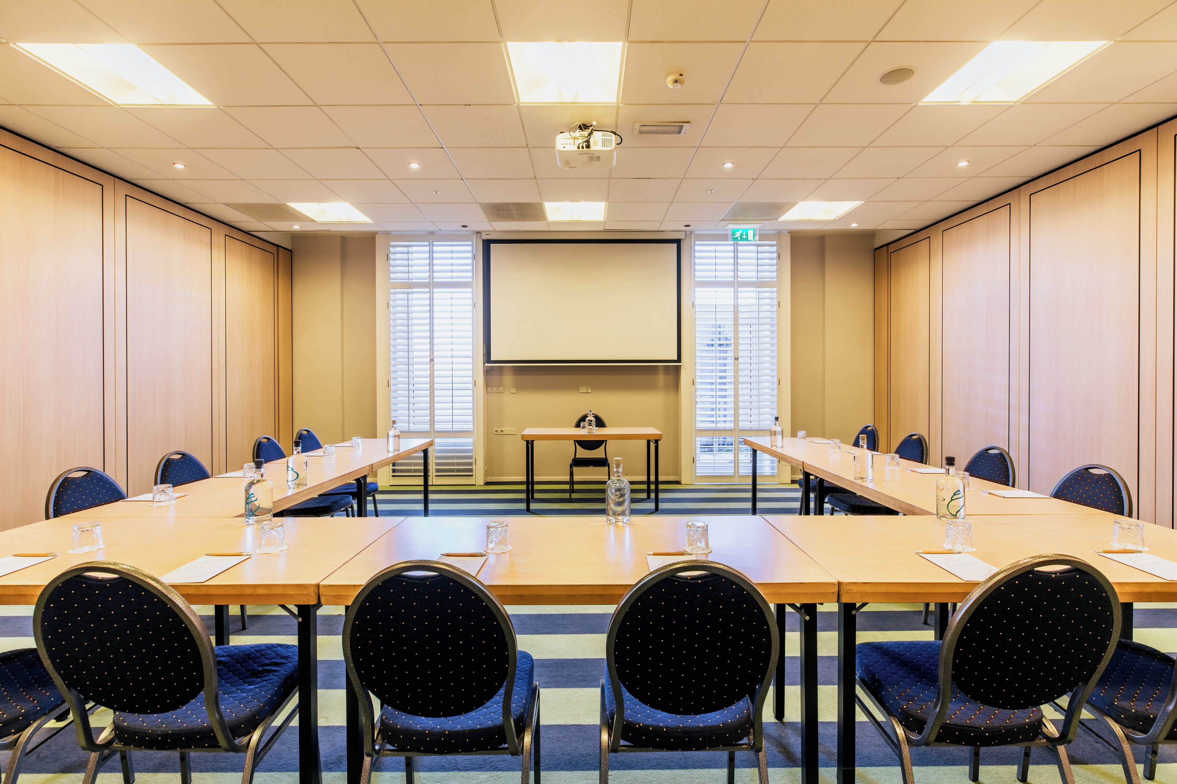 Een vergaderzaal met een lange tafel, stoelen, een projectiescherm en een presentatietafel.