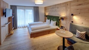 Quarto de hotel moderno com piso de madeira, cama com roupa de cama branca, cabeceira verde, sofá cinza, pequena mesa e televisão fixada na parede.