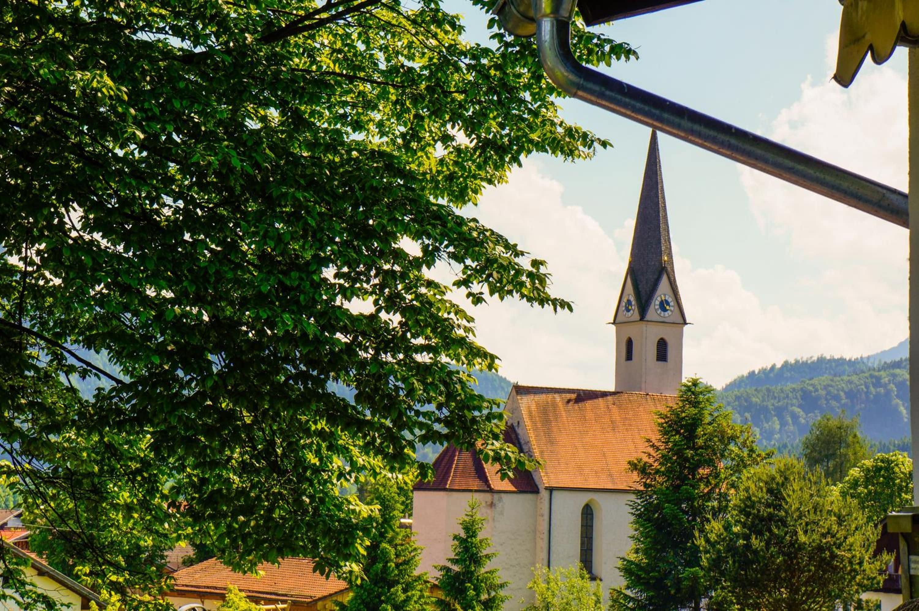 En kirke med et spidsfindigt spir og et klokketårn, omgivet af træer og et bjerg i baggrunden.