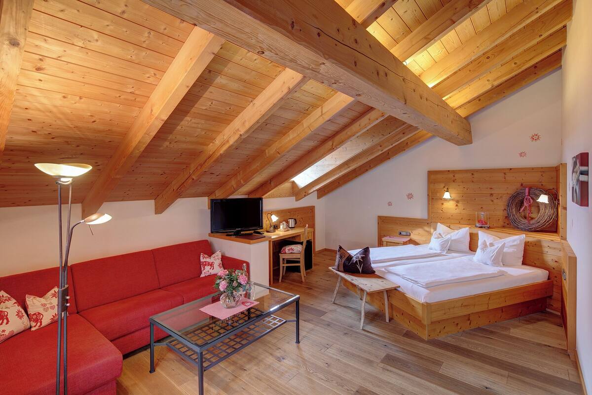 Ein gemütliches Zimmer mit Holzbalkendecke, einer Couch, einem Bett, einem Schreibtisch und einem Fernseher.
