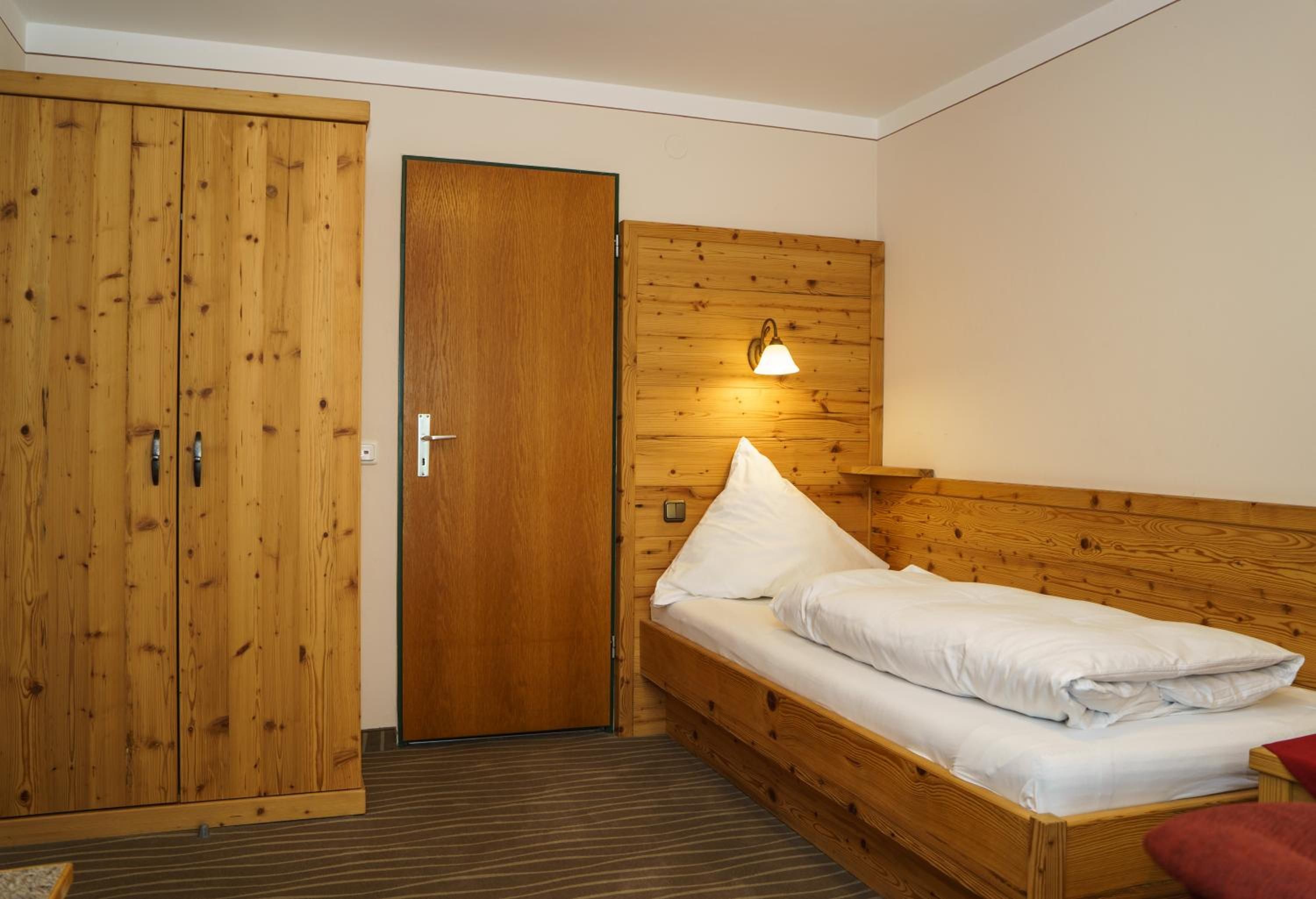 A small room with a wooden wardrobe, a bed with white bedding, and a wall-mounted light fixture.