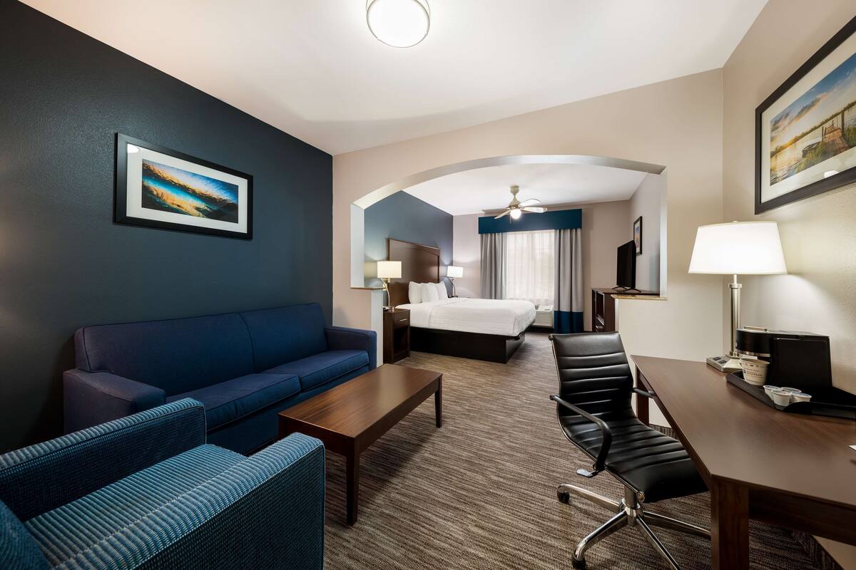 Habitación de hotel con sofá azul, mesa de centro de madera, escritorio con lámpara, cama con cabecera y un cuadro en la pared.