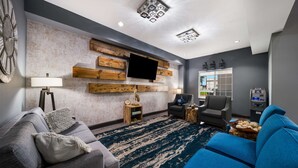 A modern living room with a grey sofa, a blue armchair, a wooden accent wall, a mounted TV, and a small side table with a lamp. - Best Western Plus Lake Dallas Inn & Suites (Lake Dallas)