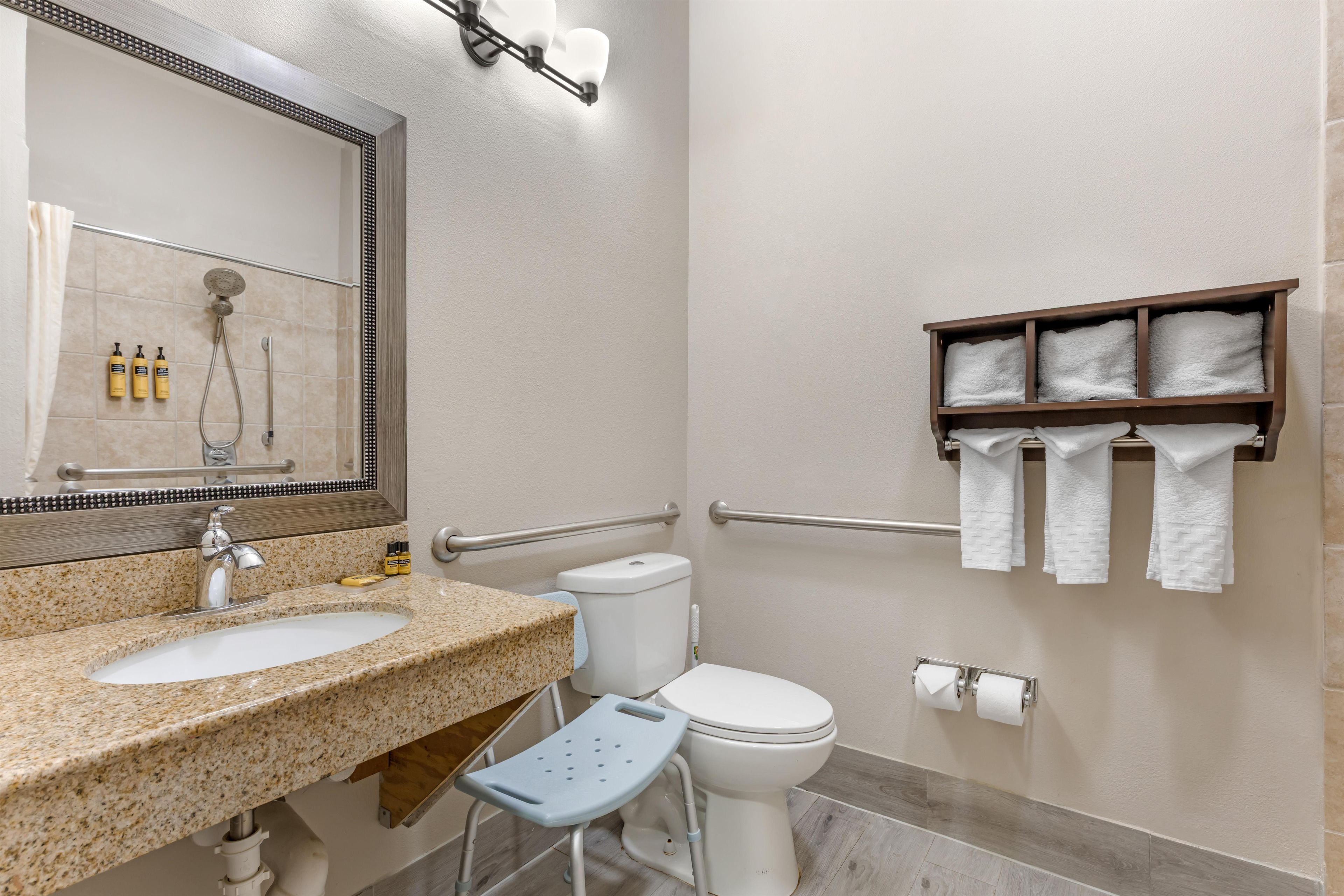 A bathroom with a granite countertop sink, a toilet, a shower with a handheld showerhead, a grab bar, and a towel rack with towels.