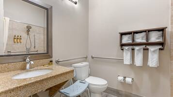 A bathroom with a granite countertop sink, a toilet, a shower with a handheld showerhead, a grab bar, and a towel rack with towels.