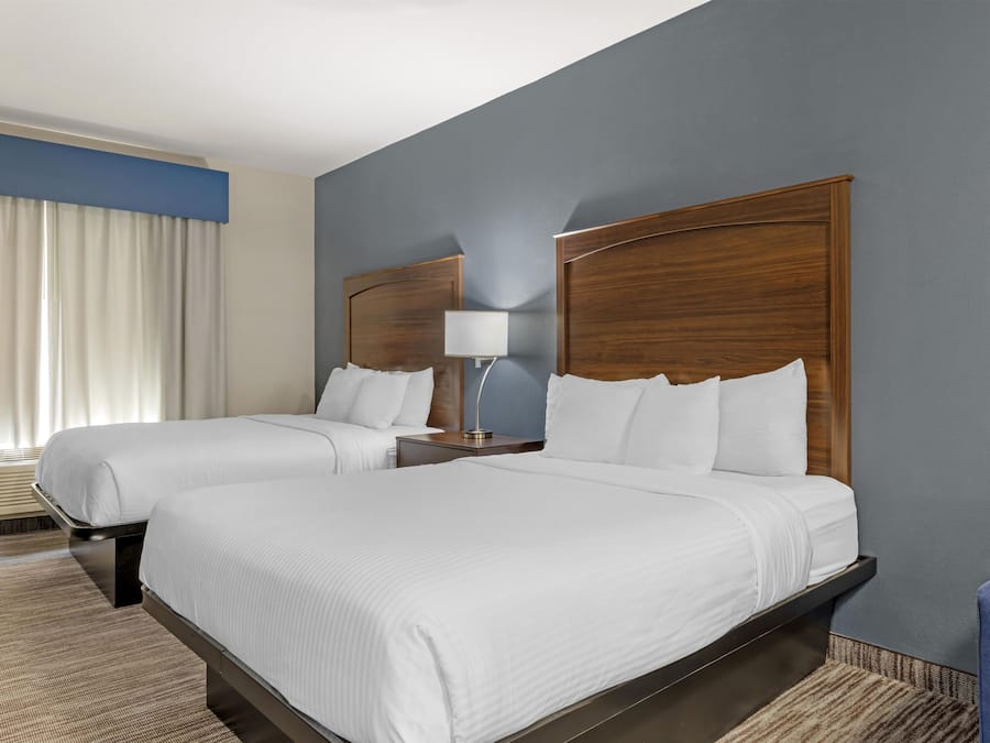 A hotel room with two beds, a nightstand, a lamp, and a large window with curtains.