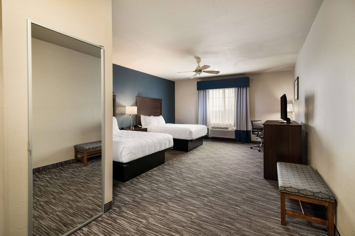 Habitación de hotel con dos camas, un escritorio, una silla, un banco, ventilador de techo y una ventana con cortinas.