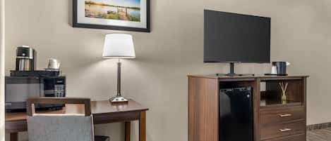 A hotel room with a bed, a desk with a lamp, a television, a coffee maker, and a minibar.
