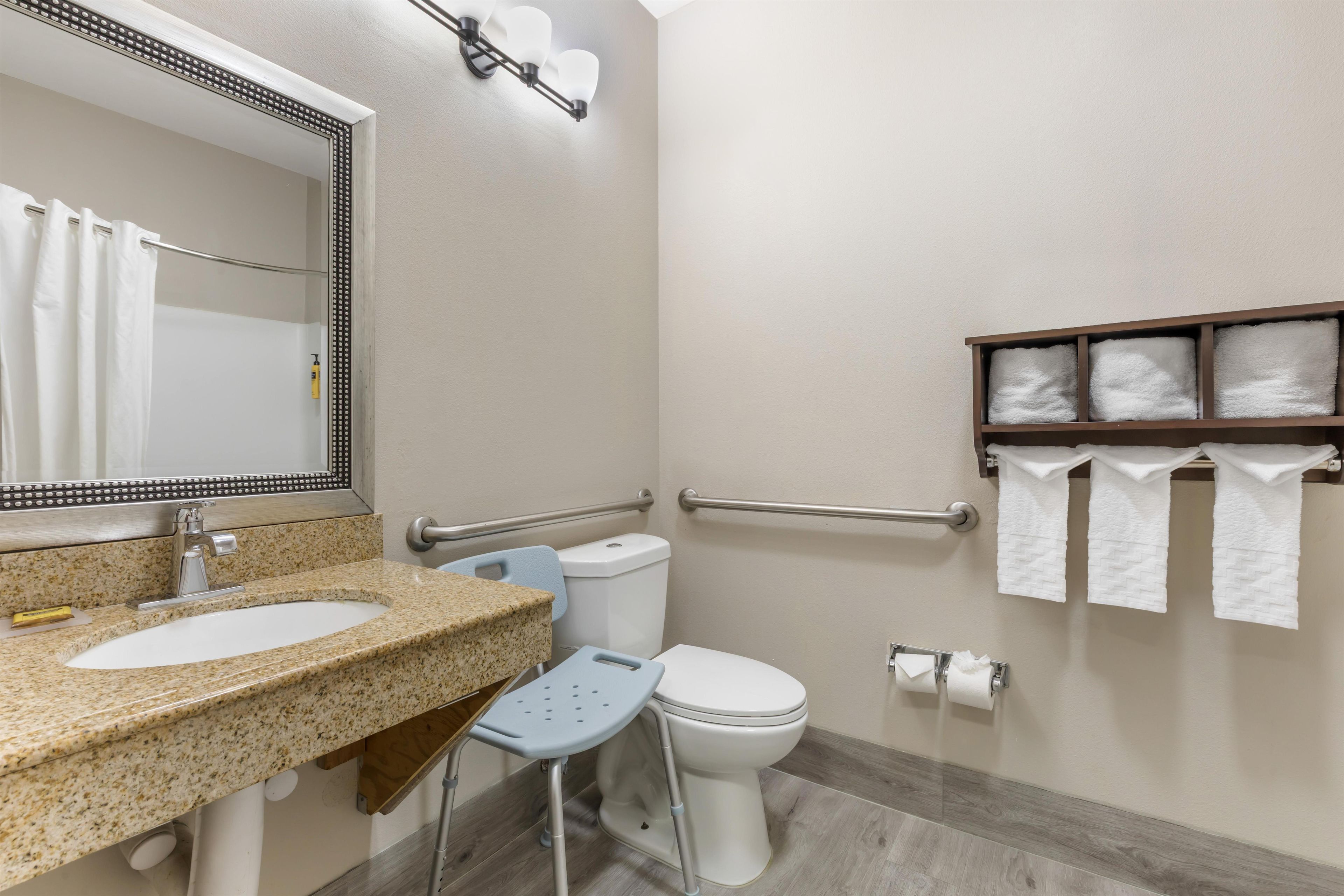 A bathroom with a granite countertop sink, a mirror with a decorative frame, a toilet, a grab bar, and a shelf with towels.