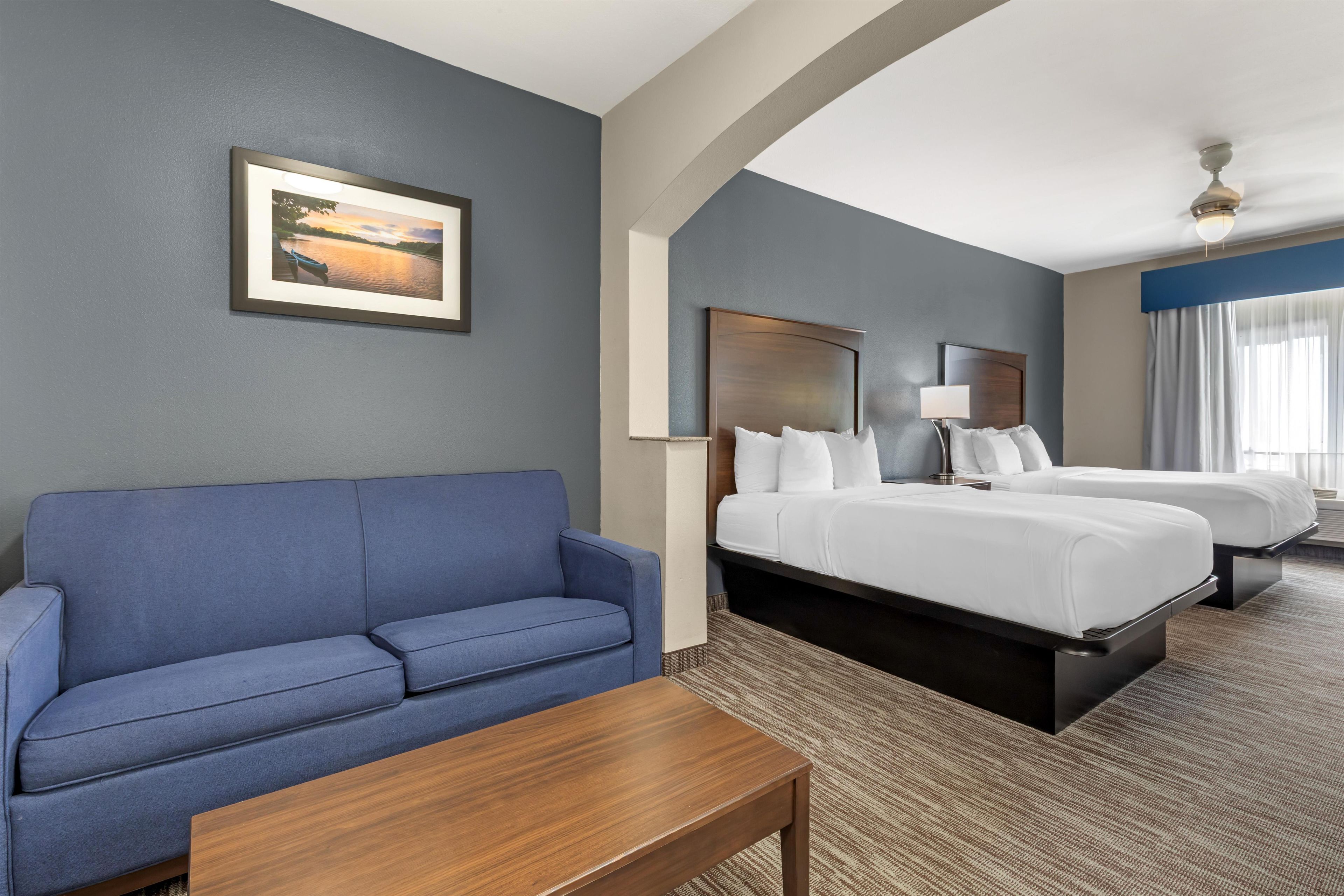 A hotel room with a blue sofa, a wooden coffee table, two beds with white linens, a framed picture on the wall, and a ceiling fan.
